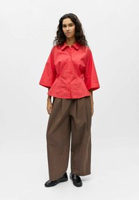 Femme debout portant une chemise rouge boutonnée à manches larges, un pantalon marron ample et des chaussures noires sans lacets sur un fond blanc.