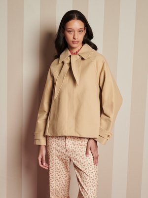 Una mujer con cabello largo y oscuro lleva una chaqueta beige con un gran cuello y un lazo, y pantalones crema con un pequeño estampado floral rosa, frente a una pared de rayas.