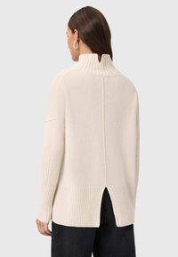 Jasna kremowa sweter z wysokim kołnierzem, z prążkowaną fakturą i szczegółem w postaci rozcięcia na dole. Posiada obniżone ramiona i miękki, przytulny wygląd.