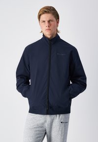 Navy athletic jacket with a high collar, full zipper, and elastic cuffs. Features a subtle Champion logo on the chest and a smooth texture.