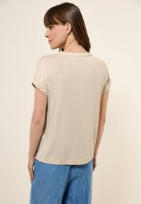 Béžové tričko s krátkými rukávy, hladký materiál a volný střih, pohled zezadu. V kombinaci s modrými denimu kalhotami.