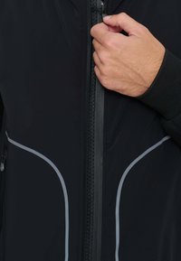 Giacca nera in tessuto liscio, con dettagli grigi e cerniera integrale. Polsini con finitura in mesh testurizzato e design aderente.