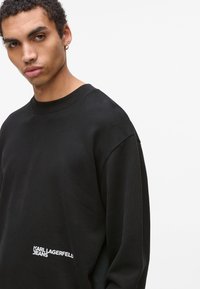 Jauns vīrietis valkā melnu džemperi ar "KARL LAGERFELD JEANS" logotipu tuvu apakšmalai uz vienkārša balta fona.