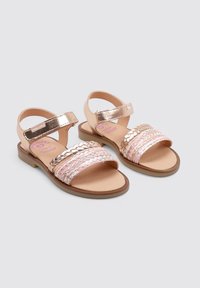 Sandalias de tiras con una base de cuero color tostado, correas metálicas doradas y detalles trenzados en rosa y plata. Suela plana con un diseño sencillo.