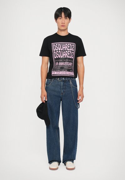 T-shirt noire avec imprimé graphique rose ; large jean en denim bleu ; chaussures blanches ; tenant une casquette noire ; fond blanc uni.