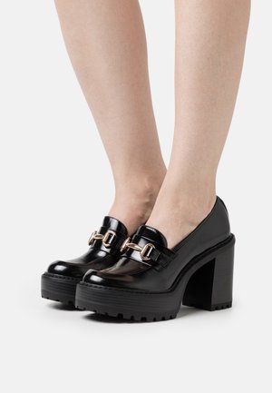 Chaussures à plateforme en cuir verni noir avec un talon épais, dotées d'accents en boucle dorée et d'une semelle en caoutchouc texturé.