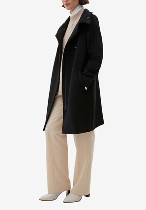 Kobieta ubrana w czarny, sięgający do połowy długości płaszcz, na beżowe spodnie o szerokich nogawkach, kremowy golf i białe botki na obcasie, stoi z rękami w kieszeniach.