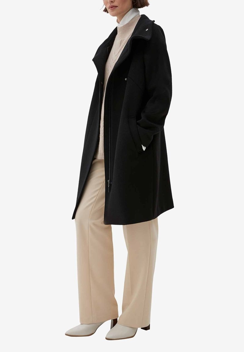 Femme portant un manteau noir mi-long sur un pantalon large beige, un pull à col roulé crème et des bottines blanches à talons, debout les mains dans les poches.