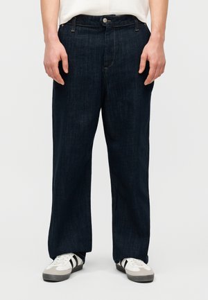 Άτομο που φοράει χαλαρά σκούρα μπλε τζιν και άσπρα αθλητικά παπούτσια με μαύρες ρίγες, στέκεται μπροστά σε μια απλή επιφάνεια.