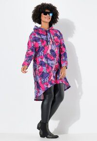 Abito con cappuccio decorato da una stampa artistica di un volto rosa e viola, con un retro più lungo, abbinato a leggins neri lucidi e stivaletti con borchie.