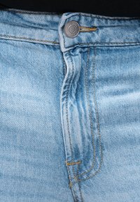 Artimas šviesiai mėlynos džinsinės medžiagos džinsų vaizdas su matomu mygtuku, užtrauktuko siūle ir oranžine siūle ant juodos liemens juostos.