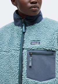 Κοντινό πλάνο ενός ατόμου που φοράει ανοιχτό μπλε μάλλινο μπουφάν με τσεπάκι στο στήθος με φερμουάρ και λογότυπο Patagonia μπροστά.