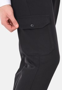 Pantaloni cargo neri con tasca applicata e chiusura a bottone. Il tessuto ha una consistenza liscia e un design aderente.