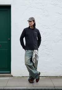 Camisa negra de manga larga, jeans azules claros de pierna ancha, gorra gris, pañuelo blanco con estampado atado a la cintura, zapatos marrones, de pie junto a una puerta verde.