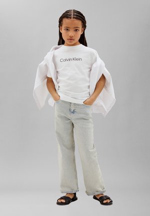 Παιδί με κοτσίδες, φοράει λευκό T-shirt Calvin Klein, ανοιχτόχρωμο τζιν, λευκό πουκάμισο κρεμασμένο στους ώμους και μαύρα σανδάλια, στέκεται με τα χέρια στις τσέπες.