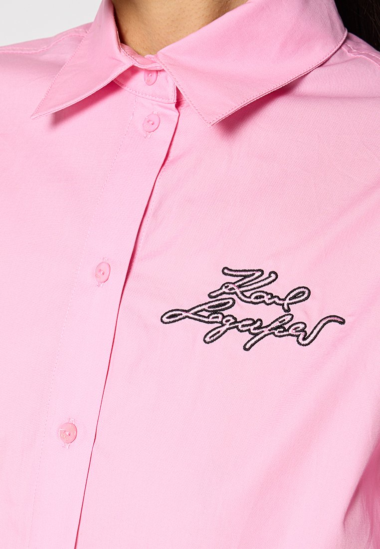 Chemise col coloris rose pâle en tissu lisse avec un plastron de boutons. Présente un texte noir brodé sur la zone gauche de la poitrine.