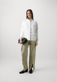 Doudoune blanche avec un col montant et des coutures horizontales, associée à un pantalon vert olive ample avec des zips et des poches.