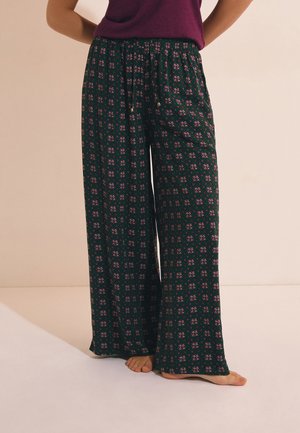 Persona que lleva pantalones verde oscuro de pierna ancha y flojos con un pequeño estampado floral púrpura y una blusa púrpura, de pie descalza sobre un suelo claro.