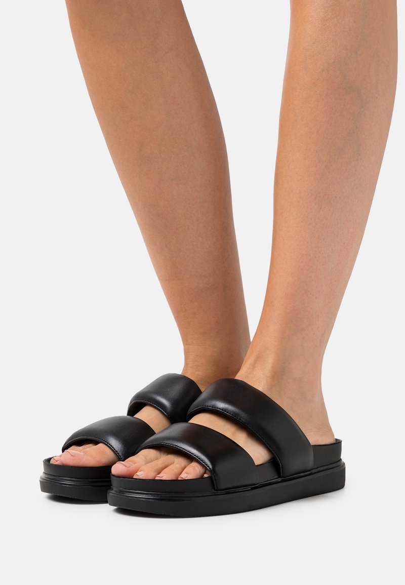Pieds portant des sandales slides noires avec deux larges brides matelassées et une semelle épaisse sur un fond clair uni.