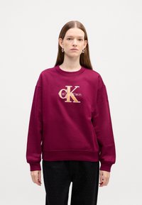 Bordový sveter s okrúhlym výstrihom a spustenými ramenami. Obsahuje vyšívané logo "Calvin Klein" v zlatej farbe a textúrovaný povrch.