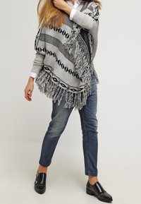 Osoba ubrana w szary frędzlony wzorzysty ponczo, białą koszulę, niebieskie dżinsy i czarne skórzane buty wsuwane, stojąca na jednolitym tle.