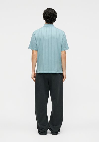Άνδρας όρθιος με την πλάτη προς τον θεατή, φοράει ανοιχτό μπλε κοντομάνικο πουκάμισο με κάθετες ρίγες και χαλαρό μαύρο παντελόνι, μπροστά σε μονόχρωμο φόντο.