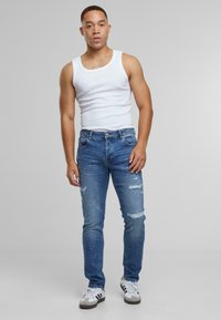 Noor mees, kes kannab valget ilma varrukat särki, rebenenud siniseid teksaseid ja valgeid tossusid, seistes ühtlasel heledal hallil taustal.