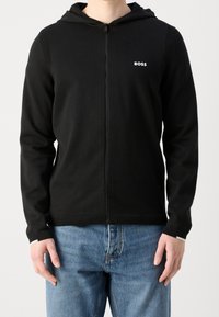 Schwarze Zip-Hoodie aus weichem Material, mit Kapuze, gerippten Akzenten und einem weißen Logo auf der linken Brust. Kombiniert mit blauen Jeans.
