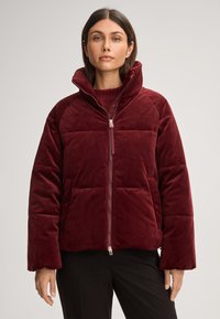 Giacca imbottita bordeaux realizzata in tessuto morbido, con colletto alto, zip frontale e design squadrato con cuciture visibili.