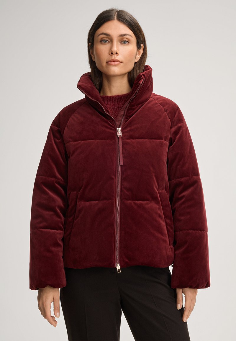 Giacca imbottita bordeaux realizzata in tessuto morbido, con colletto alto, zip frontale e design squadrato con cuciture visibili.