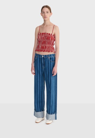 Jeune femme debout portant un haut court rouge et blanc à rayures avec des bretelles spaghetti, un jean à rayures bleues à jambes larges, et des chaussures blanches.