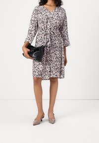 Nainen, jolla on polven mittainen abstrakti kuvioinen mekko vyötärönauhan kanssa, pitää mustaa teksturoitua laukkua ja käyttää beigejä kärjellisiä korkokenkiä.