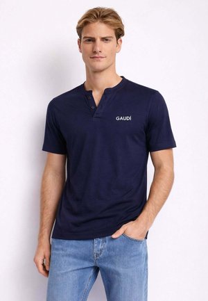 Giovane uomo con indosso una maglietta henley a maniche corte blu navy con il logo "GAUDÍ" e jeans azzurri, in piedi con una mano in tasca.