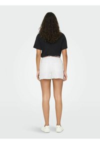 Beli denim kratke hlače z razfraknjenim robom. V kombinaciji z črno majico. Enostavna zasnova brez vzorcev. Model stoji s hrbtom ob svetli ozadju.