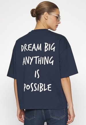 Vrouw met haar in een knot, zonnebril en een donkerblauw overhemd met de tekst "DREAM BIG ANYTHING IS POSSIBLE" op de rug.