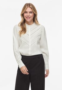 Balta ilgomis rankovėmis palaidinė su sudėtingais nėrinių detalėmis, sagomis priekyje, klostuotu dizainu ir struktūrizuotais rankogaliais, derinama su pritaikytais juodais kelnėmis.