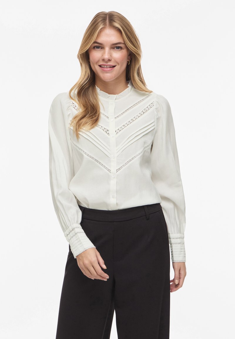 Balta ilgomis rankovėmis palaidinė su sudėtingais nėrinių detalėmis, sagomis priekyje, klostuotu dizainu ir struktūrizuotais rankogaliais, derinama su pritaikytais juodais kelnėmis.