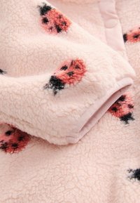 Mäkká ružová fleece tkanina s malými kvetinovými vzormi v červenej a čiernej farbe. Má hladký lem a detailné gombíky pozdĺž okraja.