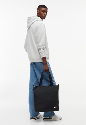 Joven con sudadera con capucha gris claro y jeans azules, sosteniendo una bolsa tote negra de Tommy Jeans por las asas, sobre un fondo blanco liso.