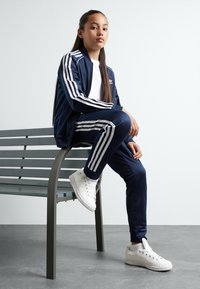 Mornarska Adidas trenirka z belimi črtami, jakna z zadrgo, oprijete hlače in bele superge. Sedi na sivi klopi ob svetli steni.
