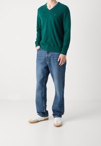 Maglione verde a scollo a V realizzato in tessuto morbido, abbinato a jeans blu e sneakers bianche con suola beige. Presenta un piccolo logo sul petto.