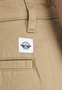 Khaki pants with a smooth texture, featuring a white tag with black logo and wings. Includes a side pocket with a straight stitch detail.