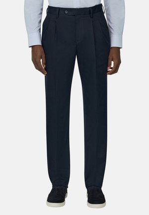 Mężczyzna ubrany w czarne garniturowe spodnie, jasnoniebieską koszulę oraz czarne wsuwane buty z białymi podeszwami, stojący na jednolitym tle.