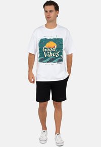 Wit katoenen T-shirt met een groene en oranje afbeelding met golven en de tekst "Good Vibes", gecombineerd met een zwarte korte broek en witte sneakers.