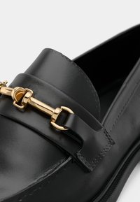 Les mocassins en cuir noir présentent un accent en chaîne dorée polie à l'avant et une texture lisse, mettant en valeur l'artisanat et les éléments de design.