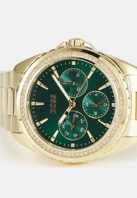 Montre-bracelet dorée avec un cadran vert, trois sous-cadrans et une lunette incrustée de cristaux. Présente des index horaires en argent et un bracelet en métal.