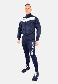  trening de marină dintr-un material neted, cu un accent alb pe jachetă și branding mare "GIVOVA" pe pantaloni. Adidașii atletici completează look-ul.