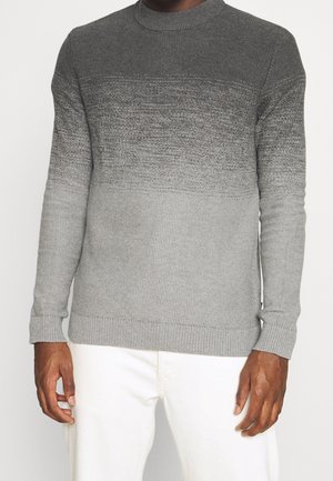 Homme portant un pull en tricot à manches longues avec un dégradé du gris foncé au gris clair et un pantalon blanc, debout sur un fond blanc.