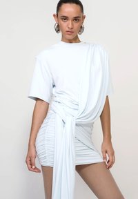 Femeie îmbrăcată într-o rochie mini cu pliuri de culoare albastru deschis, cu material drapat și cercei mari argintii, stând pe un fundal simplu.