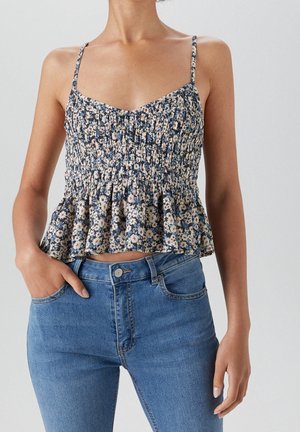 Vrouw draagt een mouwloze, met bloemenmotief gesmokte top met spaghettibandjes en blauwe high-waisted jeans, staand met één hand in de zak.